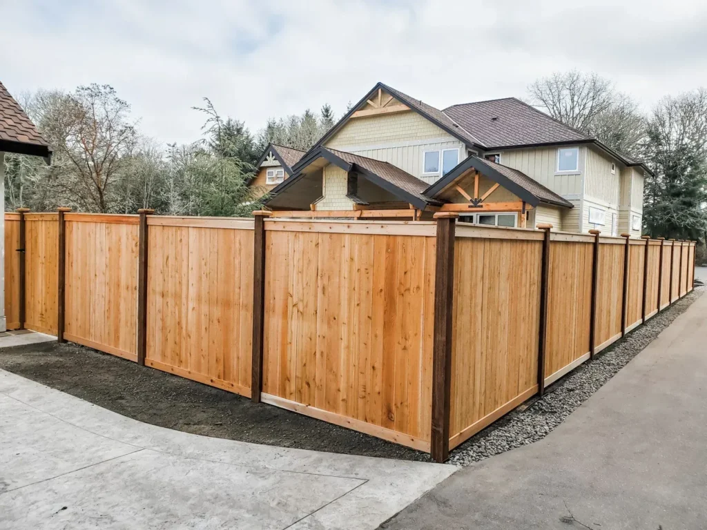 A newly built cedar privacy fence with capped posts wrapping a property corner, gravel on one side and a stamped-concrete driveway on the other, with a large craftsman house behind